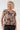 A woman with curly blonde hair wearing a short-sleeve scoop-neck top with an orange and gray abstract circular pattern on a dark teal background, 