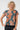 A smiling blonde woman wearing a short-sleeve blouse with a teal blue base and a large orange and gray sunburst floral pattern, 