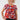 A smiling woman with curly blonde hair wearing a red short-sleeve top with a colorful abstract geometric pattern, 