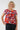 A smiling woman with curly blonde hair wearing a red short-sleeve top with a colorful abstract geometric pattern, 