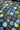 A close-up of a fabric surface with an allover floral or pom-pom print in blue, light blue, green, white, and gray on a dark charcoal background.